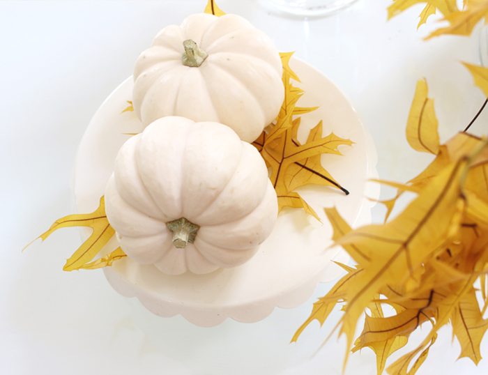 two white mini pumpkins on a cake stand with yellow leaves