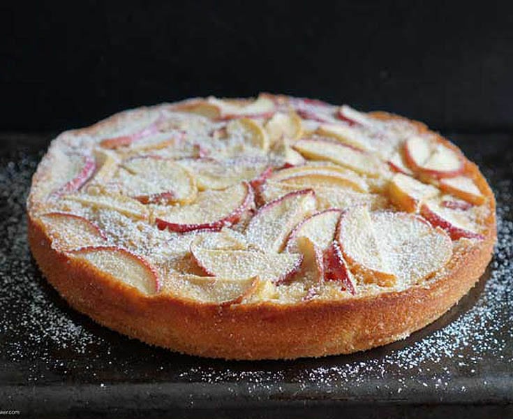 whole apple tart with powdered sugar sprinkled on top. Tart is placed on a black table top