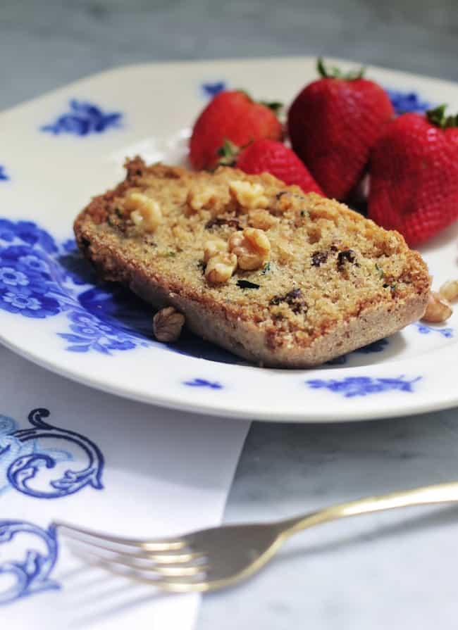 A slice of nutty bread with visible walnuts sits on a white plate with blue floral designs, accompanied by three whole strawberries—an inviting scene that hints at the best recipes for breakfast. A silver fork and patterned napkin rest nearby on the marble surface.