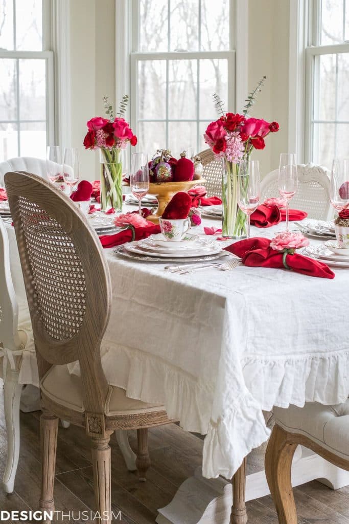 Elegant Valentine's Day dinner table set with a white ruffled tablecloth, red napkins, and crystal glassware. Red and pink floral centerpieces adorn the scene in glass vases. Intricate wooden-backed chairs and large windows complete this romantic setting.