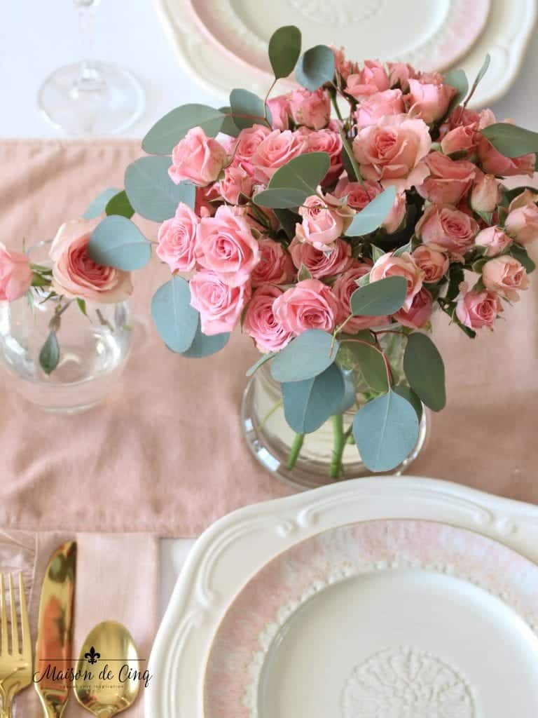 A bouquet of pink roses with eucalyptus leaves in a glass vase adorns the Valentine's Day dinner table, complemented by a pink runner. The table is elegantly set with white and pink floral-patterned plates and gold utensils, creating a romantic ambiance.