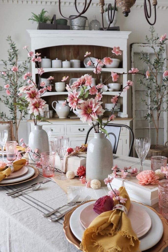 A Valentine's Day dinner table is elegantly set with pink flowers in white vases, surrounded by crystal glasses, pink plates, yellow napkins, and silver cutlery. The background features a white hutch with white dishes and decorative plants.