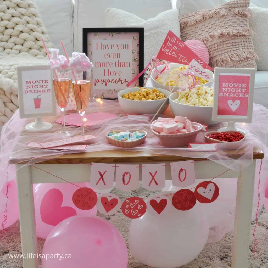 A Valentine's Day dinner table setup features pink and red decorations, popcorn, candies, drinks, and a frame with romantic messages. Balloons and a "XOXO" garland enhance the festive atmosphere.