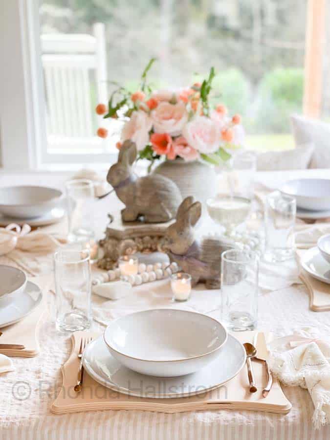 Elegant Valentine's Day dinner table setting with white and blush tones. Features stacked bunny figurines, a pink and white floral centerpiece, white plates, clear glasses, and gold cutlery on a soft-striped tablecloth. Candles add a warm glow.