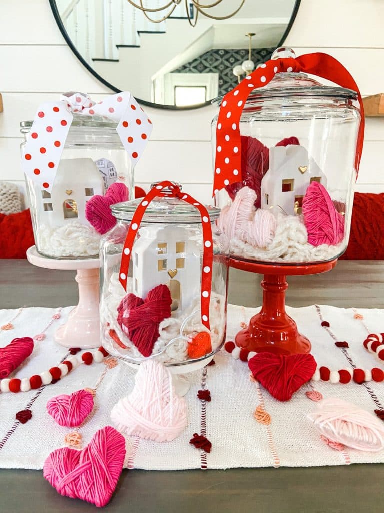 Three decorative glass jars filled with red and pink yarn, each topped with a bow, stand on cake stands. Perfect for a Valentine's Day dinner table, yarn hearts and small houses nestle inside the jars. A table runner with yarn hearts and garland adorns the setup, while a mirror reflects a staircase wall.