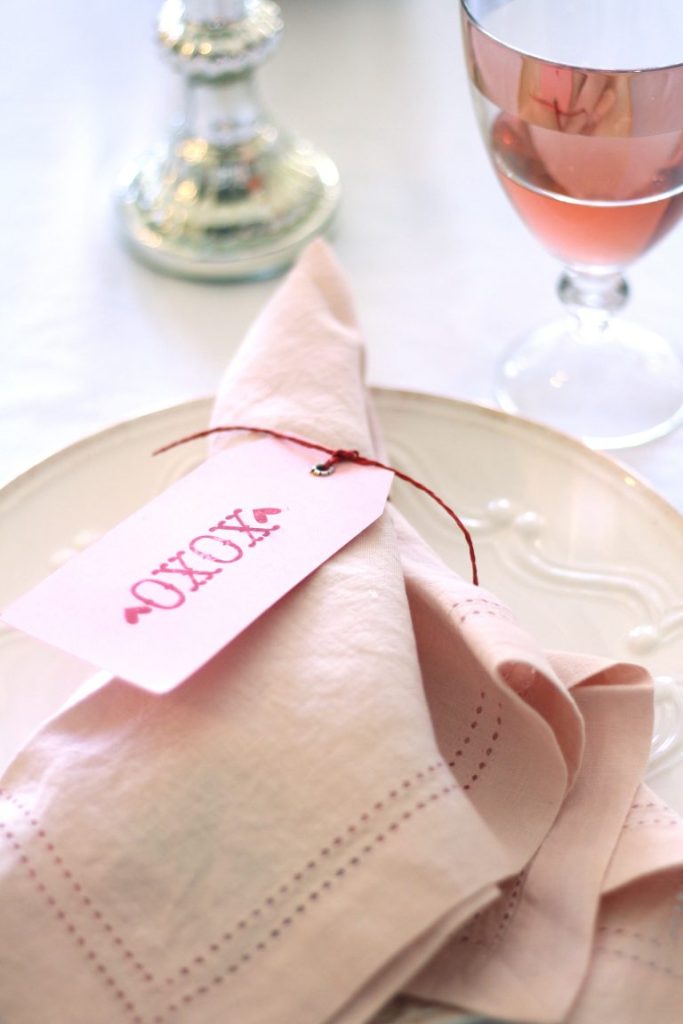 A pale pink napkin folded neatly on a white plate with a tag reading "XOXO" attached by red string hints at a romantic Valentine's Day dinner table. A glass of pink liquid and a shiny candlestick adorn the softly lit setting in the background.