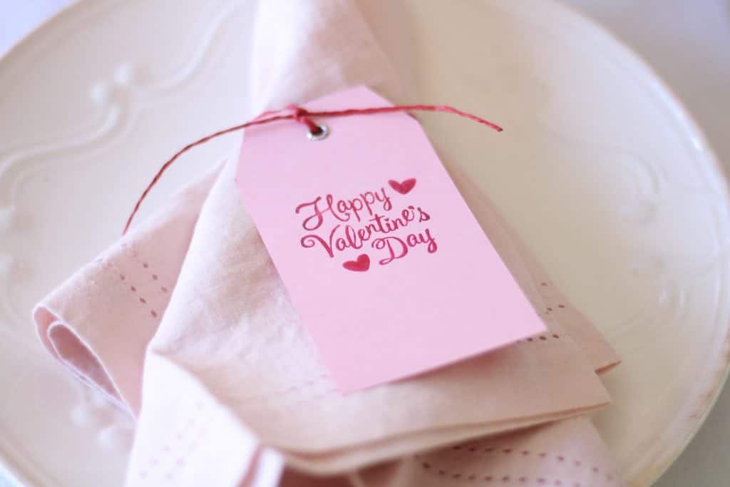 A pink napkin graces the white plate, tied with a red string and a pink tag reading "Happy Valentine's Day" in red cursive. Small heart illustrations adorn it, setting the perfect touch for your Valentine's Day dinner table.