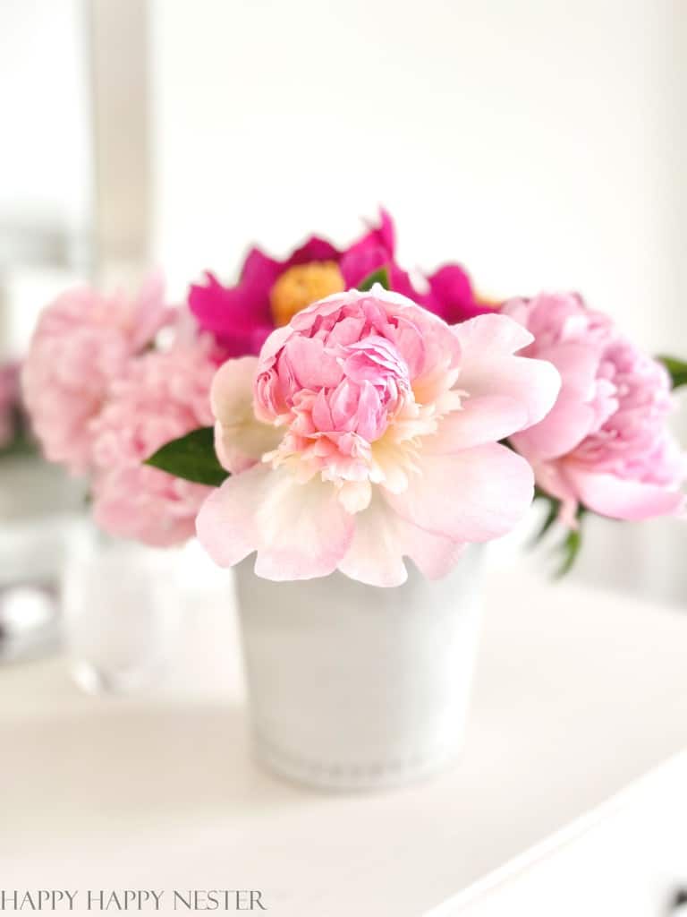 A white vase with pink peonies in it. The vase and flowers are sitting on a white table.