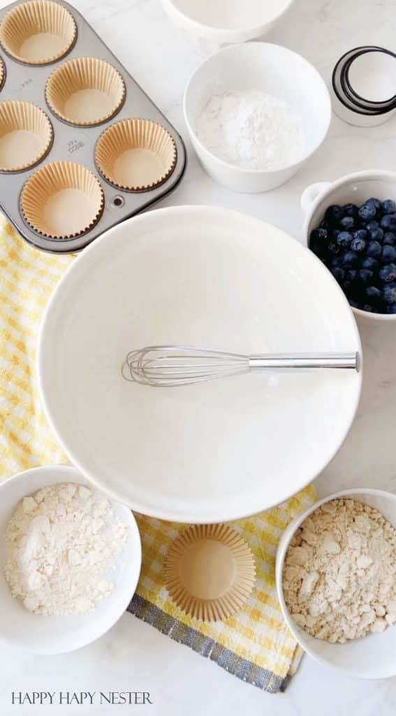 a white bowl with a whisk in it with all the almond flour blueberry muffins ingredients next to it.