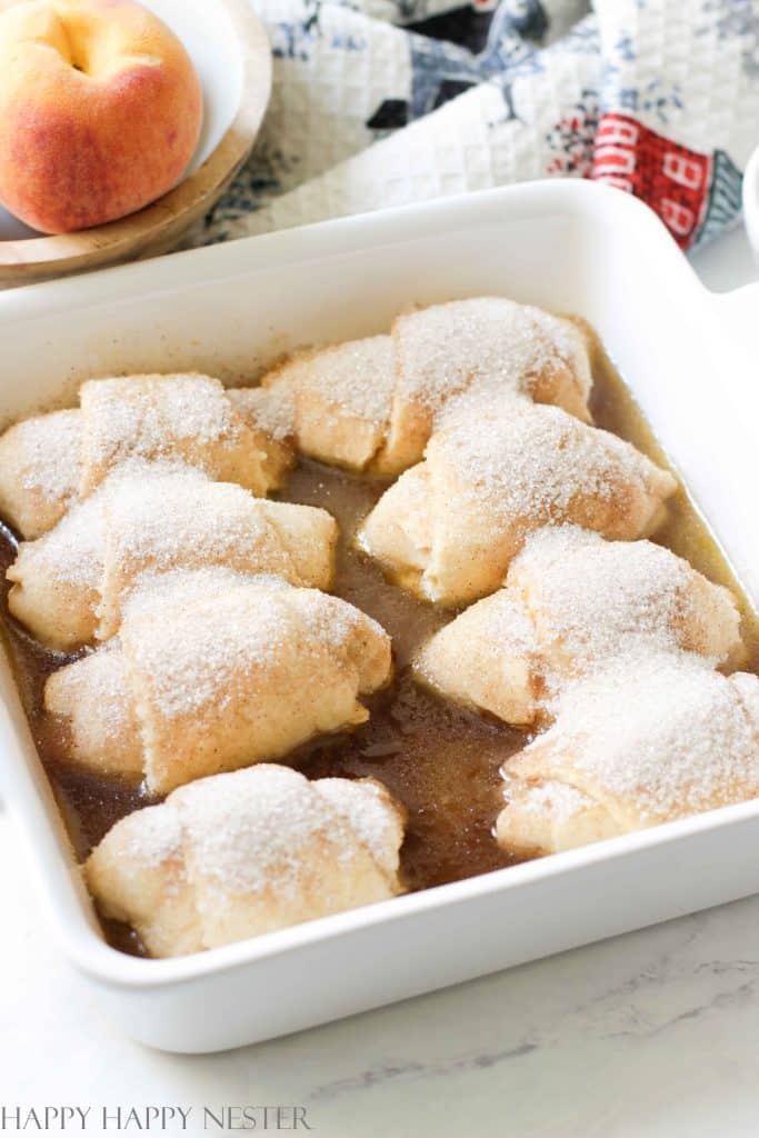 8 peach dumplings rolled up with crescent roll dough are lined up in a white casserole dish. They are surrounded by a caramel sauce that is ready to be baked. 
