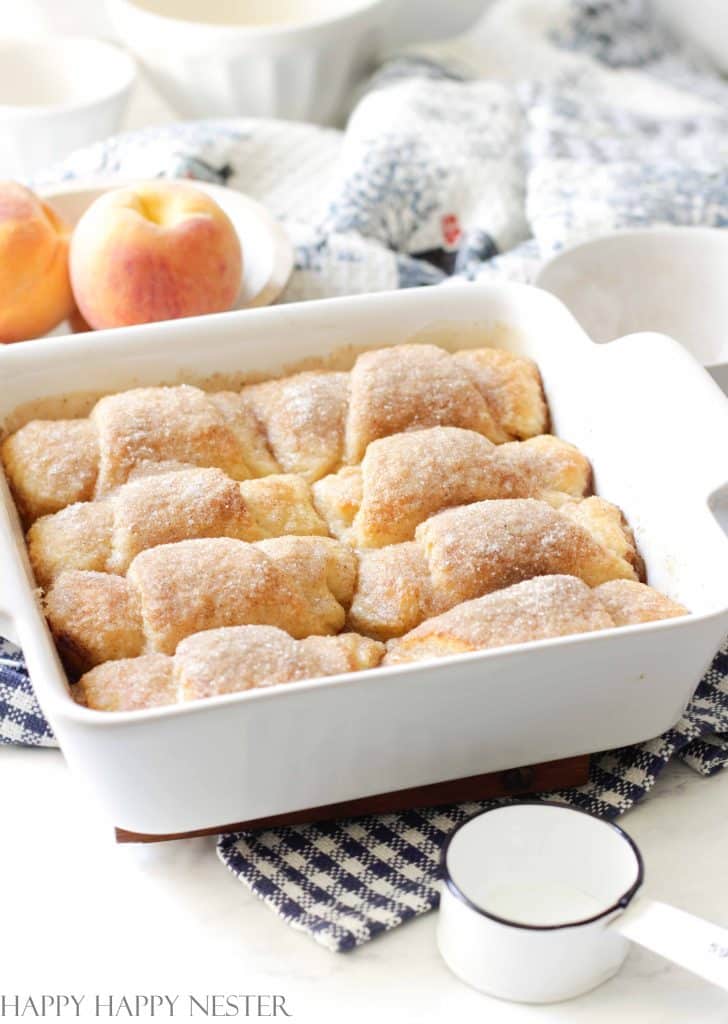 Crescent rolls sitting in a caramel sauce all in a white casserole dish. Peaches are beside the dish and a blue dish towel is surrounding the casserole dish. 
