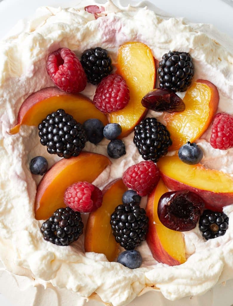 A close up of the top of a pavlova. Fruits like peach slices, raspberries, cherries and blueberries are pictured