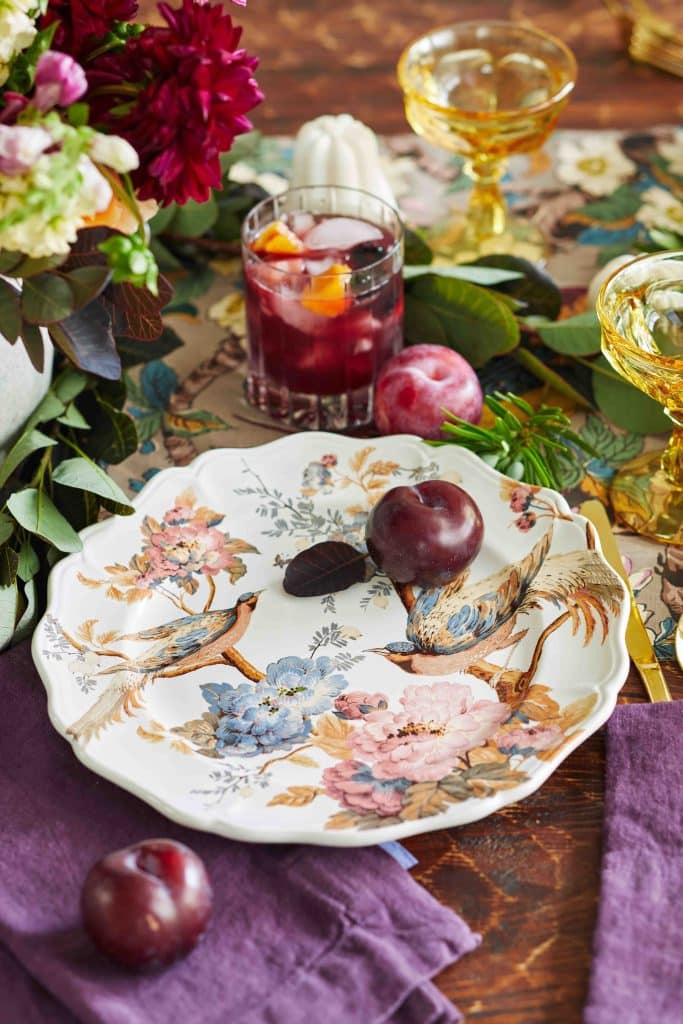 A beautiful bird themed dinner plate with a purple plum on the plate. Off to the side is a dark sangria in a crystal glass. 