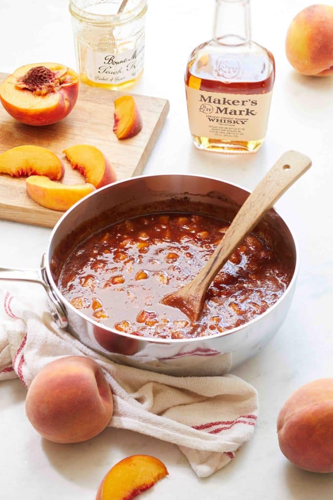 Peach bourbon glaze in a pan with a wooden spoon. There is a whisky bottle and sliced peaches surrounding the glaze.