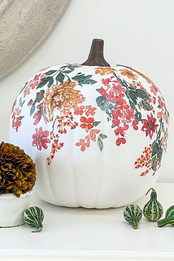 A white pumpkin decorated with colorful floral patterns in shades of orange, red, and green sits on a white surface, surrounded by small green gourds—a lovely example for pumpkin craft ideas.