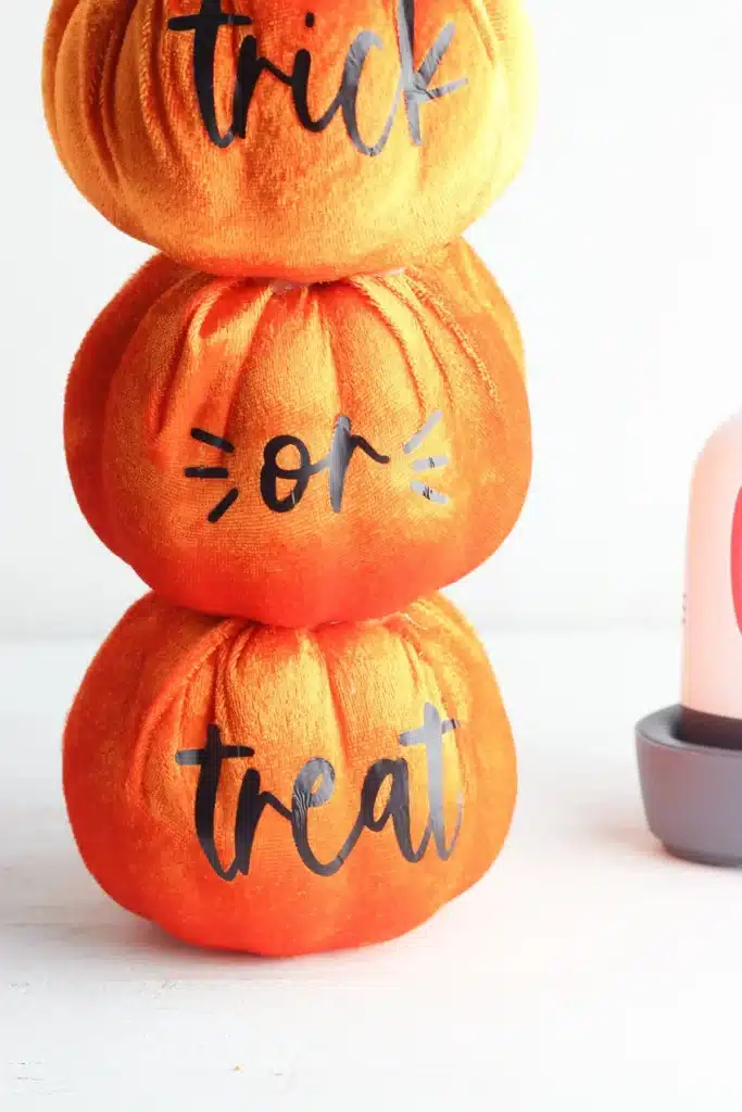 Three stacked orange fabric pumpkins with shiny black "trick or treat" lettering from top to bottom—an inspiring addition for pumpkin craft ideas. Part of a bottle and a gray container appear on the right side of the image.