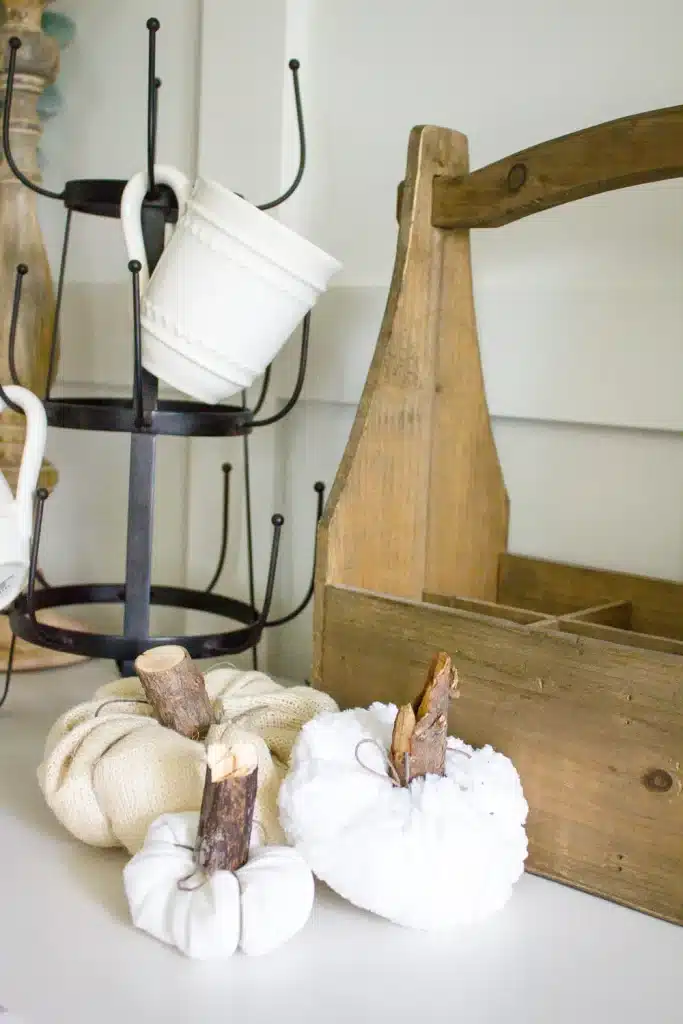 Three decorative fabric pumpkins with wood stems sit on a white surface next to a rustic wooden caddy and a black metal mug rack with white mugs—perfect inspiration for farmhouse autumn decor and pumpkin craft ideas.