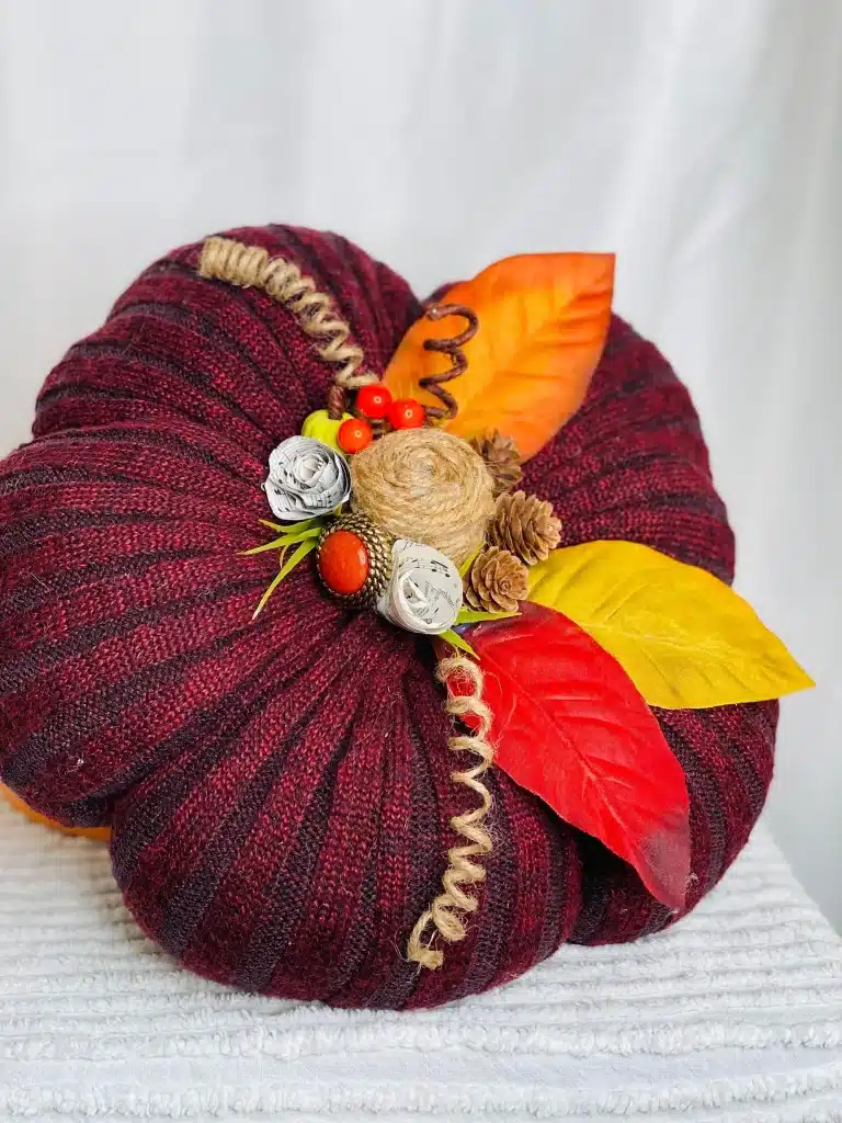 A decorative pumpkin made from deep red knit fabric, adorned with artificial autumn leaves, small pinecones, faux berries, and twine accents, offers inspiration for cozy pumpkin craft ideas on a white textured surface against a white background.