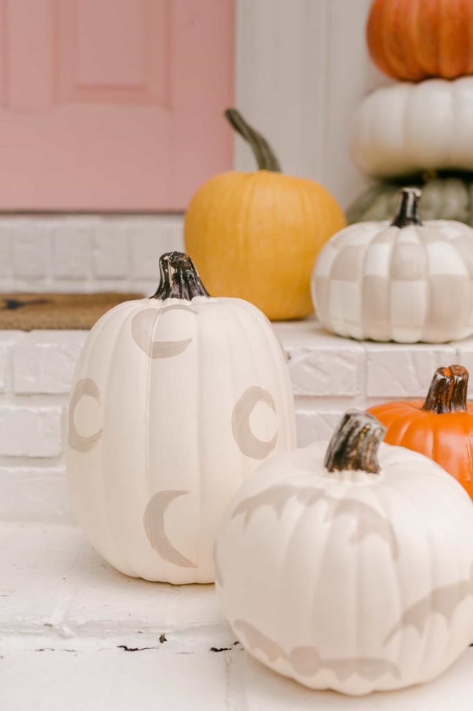 White pumpkins painted with moon shapes are displayed on white steps, alongside yellow, orange, and checkered pumpkins—a charming showcase of pumpkin craft ideas—with a pink door in the background.