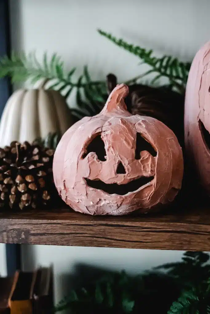A pink jack-o’-lantern with a smiling face sits on a wooden shelf, surrounded by pinecones, a white pumpkin, and green fern leaves—perfect for those looking for creative pumpkin craft ideas.