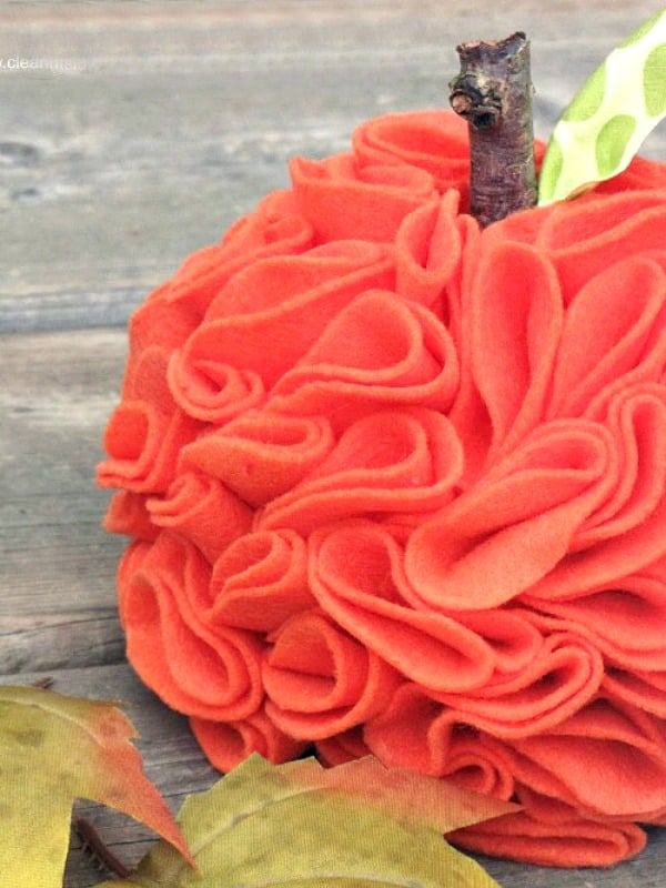 A close-up of a decorative pumpkin made from layered orange felt fabric, with a small twig stem and green fabric leaf, resting on a wooden surface next to yellow leaves—perfect inspiration for pumpkin craft ideas.