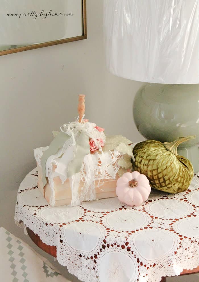 A round table with a white, lacy tablecloth displays a green lamp, two decorative pumpkins (one green and one pink) perfect for pumpkin craft ideas, and a stack of books wrapped in lace and ribbon with a wooden knob on top.