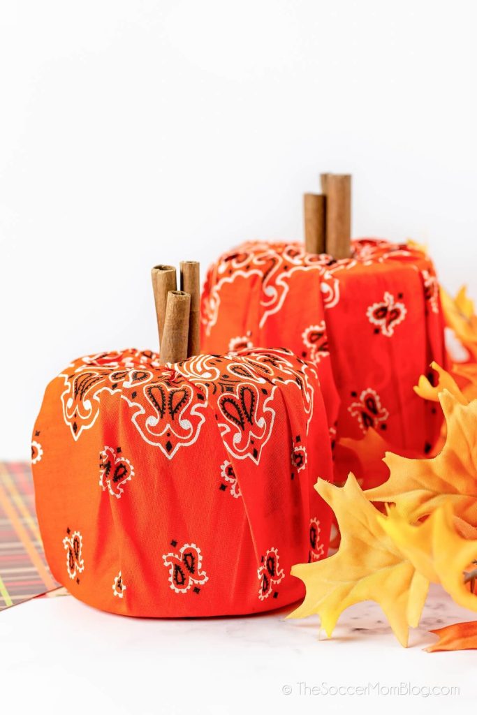 Two decorative pumpkins made from orange bandanas and cinnamon stick stems sit on a white surface, with yellow artificial autumn leaves beside them—perfect inspiration for your next pumpkin craft ideas.