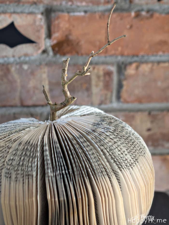 An open book with its pages folded into a round, pumpkin-like shape and a twig as a stem—an inspiring example for pumpkin craft ideas—set against a brick wall background.