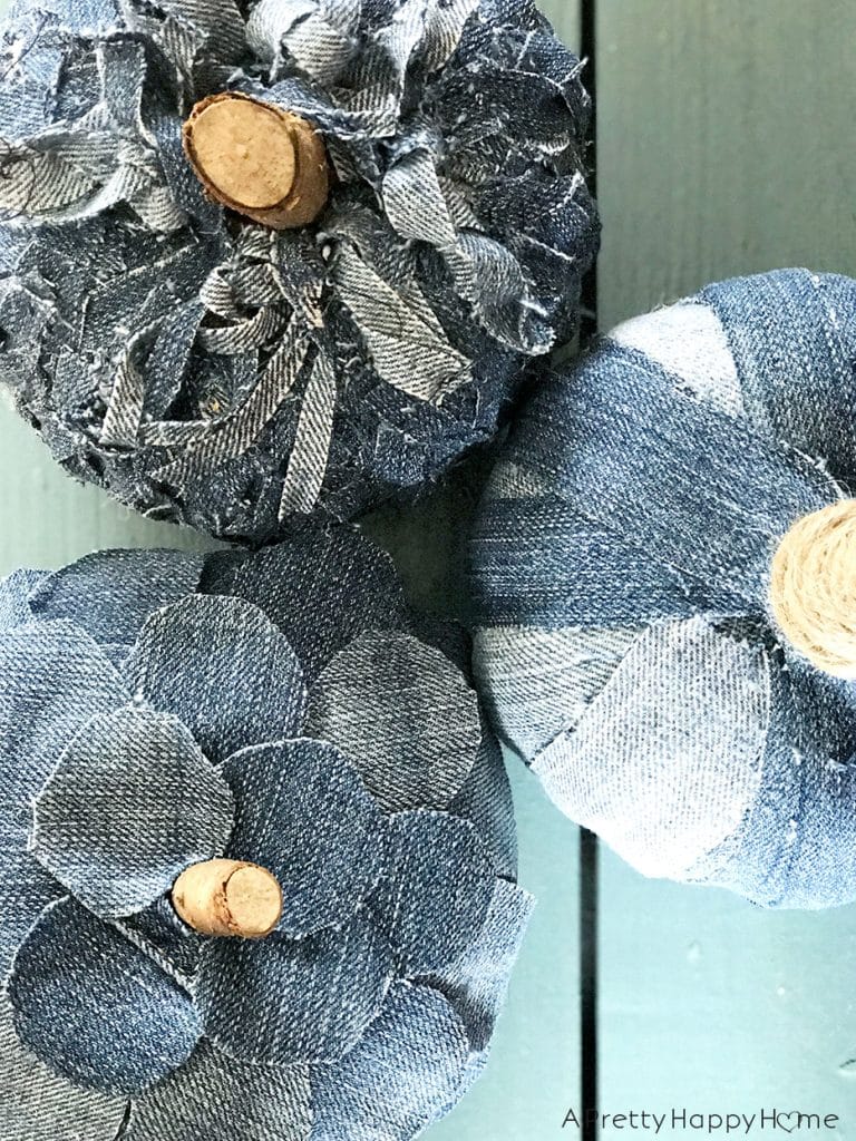 Three pumpkins made from repurposed denim fabric and wooden stems are displayed on a teal wooden surface—unique pumpkin craft ideas with petal-like segments and a striped patchwork design add rustic charm.