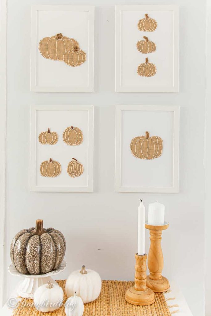 Four framed artworks featuring textured pumpkin shapes—a fun way to showcase pumpkin craft ideas—hang on a white wall above a table with a glittery pumpkin, two white pumpkins, and two wooden candle holders with white candles.