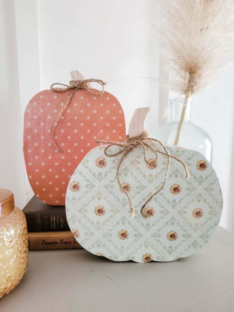 Two decorative wooden pumpkins, inspired by pumpkin craft ideas—one pink with white polka dots and the other pale blue with a floral pattern, both tied with twine bows—are displayed on a shelf next to books, a glass vase, and a candle.