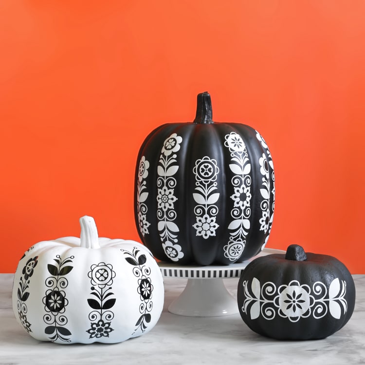 Three pumpkins, two white and one black, display intricate black and white floral folk art patterns—perfect inspiration for your next pumpkin craft ideas. They sit on a white surface against a vibrant orange background.