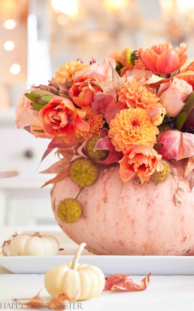 A light pink pumpkin is used as a vase, filled with vibrant orange flowers and autumn leaves—a charming example of pumpkin craft ideas—sitting on a white tray with two small white pumpkins beside it. The background is softly blurred.