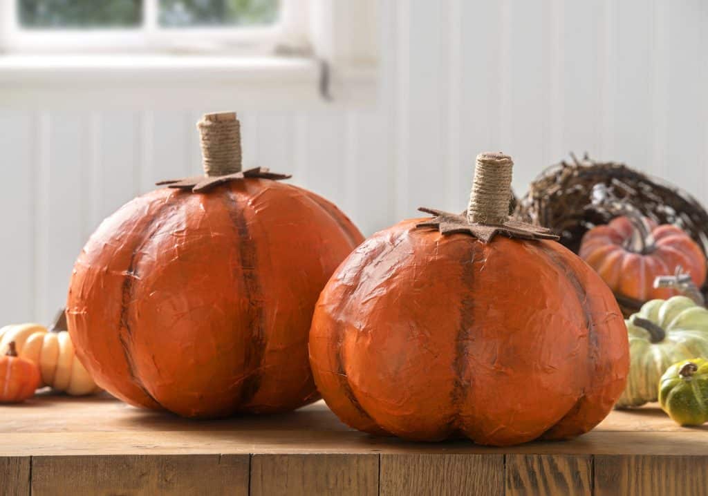 Two handcrafted paper mache pumpkins with twine stems sit on a wooden table, surrounded by other decorative gourds—perfect inspiration for pumpkin craft ideas. A white window and walls are visible in the background.