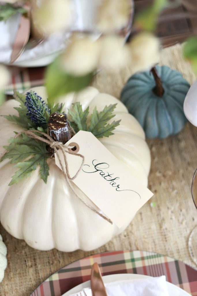 A white pumpkin decorated with greenery, twine, and a lavender sprig sits on a table—perfect for those seeking pumpkin craft ideas. A "Gather" tag hangs from the stem, accompanied by a blue pumpkin and a plaid-patterned plate.