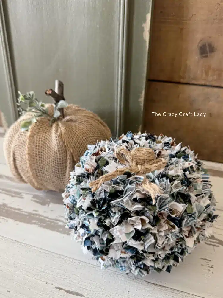 Two decorative pumpkins on a wooden surface showcase unique pumpkin craft ideas: one is covered in burlap with a twine stem, while the other is made from strips of patterned fabric tied together for a textured look.
