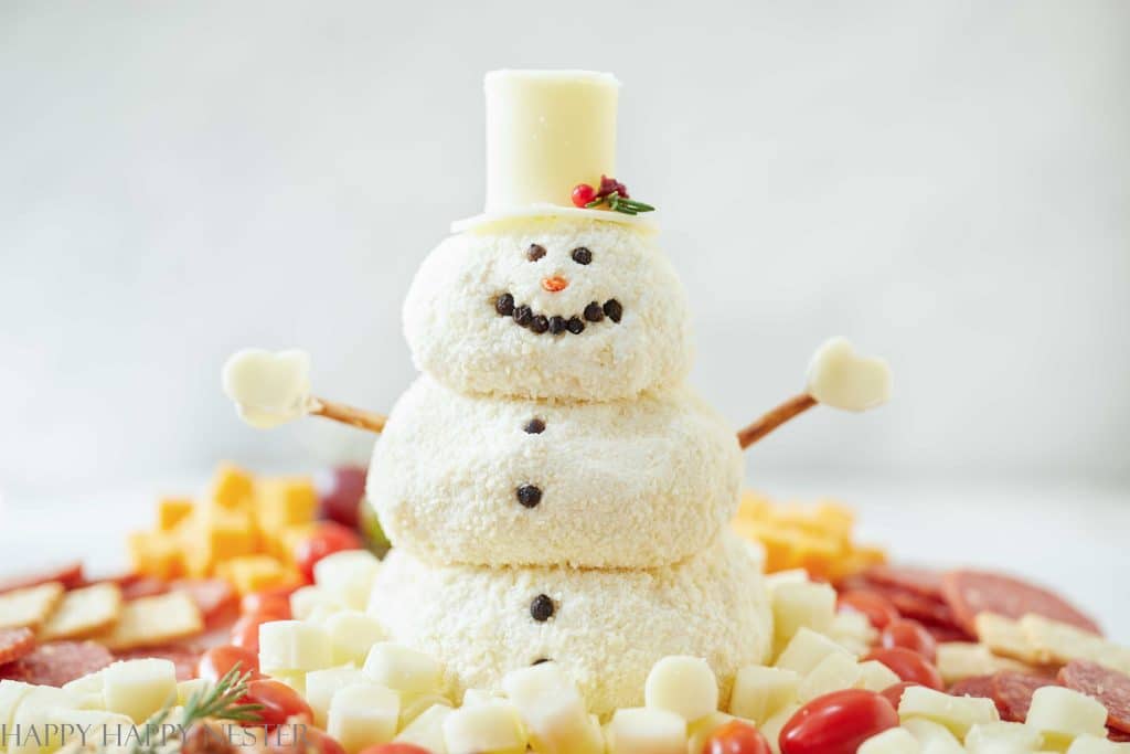 a cute snowman made out of cheese with pretzel arms and cheese mittens. He is sitting on the top of fruit and cheese charcuterie board. 