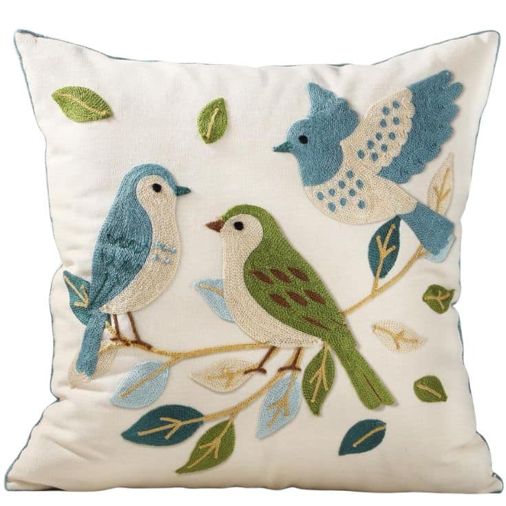 A square cream-colored springtime throw pillow featuring embroidered designs of three birds and green, blue, and beige leaves on branches. The birds are in shades of blue, green, and beige, adding a fresh nature-inspired touch.