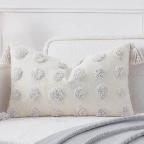 A cream-colored decorative pillow with white tassels on each corner and raised, fluffy polka dots—one of the chic amazon boho throw pillows—rests on a white sofa with a gray blanket draped over the seat.