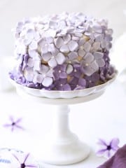 A beautifully decorated cake sits on a white pedestal. The cake is adorned with numerous light purple and lavender floral decorations, giving it a charming and elegant appearance. A purple flower lies next to the pedestal on a white surface.