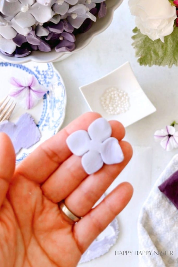 A hand holding a delicate, light purple flower-shaped fondant piece. In the background, there are various crafting supplies including a small plate with a fork and another fondant flower, a dish with small white beads, and pieces of purple and white fabric.