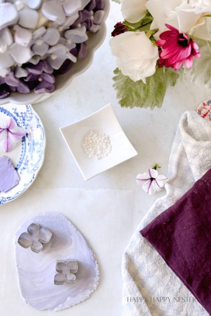 A crafting scene featuring a rolled-out piece of light purple clay with flower-shaped cutters, a bowl of small white beads, a cloth with a purple flower, and a bouquet of white and pink flowers on a table. Text at the bottom reads "HAPPY HAPPY NESTER.