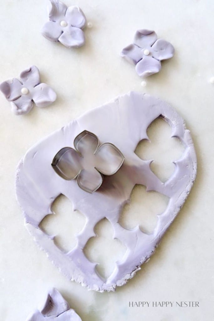 A floral-shaped cookie cutter is surrounded by cut-out flowers made from light purple fondant or clay. The cutter rests on a sheet of rolled-out purple material with several flower shapes cut out. Some completed flower pieces are set around the cutter. Text reads "HAPPY HAPPY NESTER.