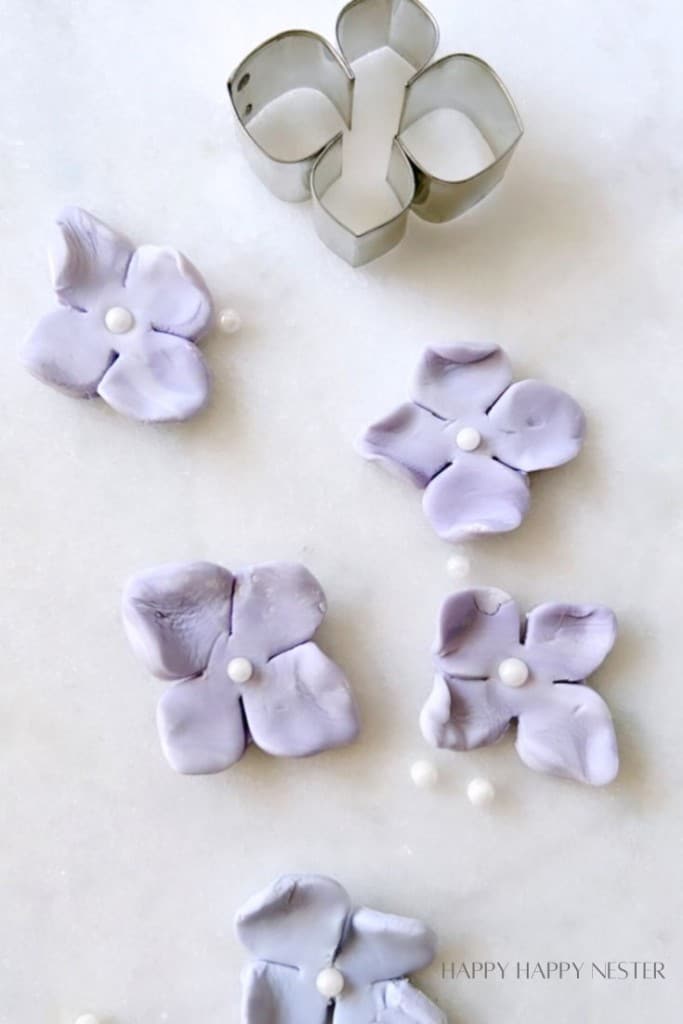A close-up of lilac fondant flower decorations on a white surface, accented with small white candy beads in the center of each flower. A edible fondant flower-shaped stencil is placed among the decorations. The words "HAPPY HAPPY NESTER" are faintly visible at the bottom right.