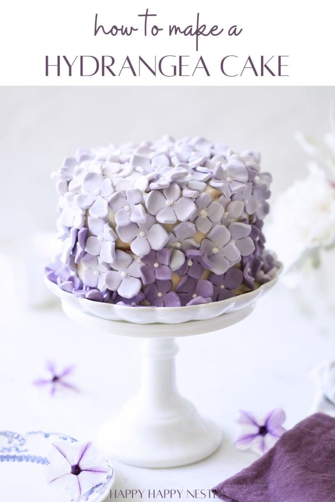 A beautifully decorated cake with purple hydrangea flowers sits on a white cake stand. The cake has detailed flower petals and a soft gradient of purple hues. The text above reads, "how to make a HYDRANGEA CAKE." Several purple flower petals and decor surround the base.
