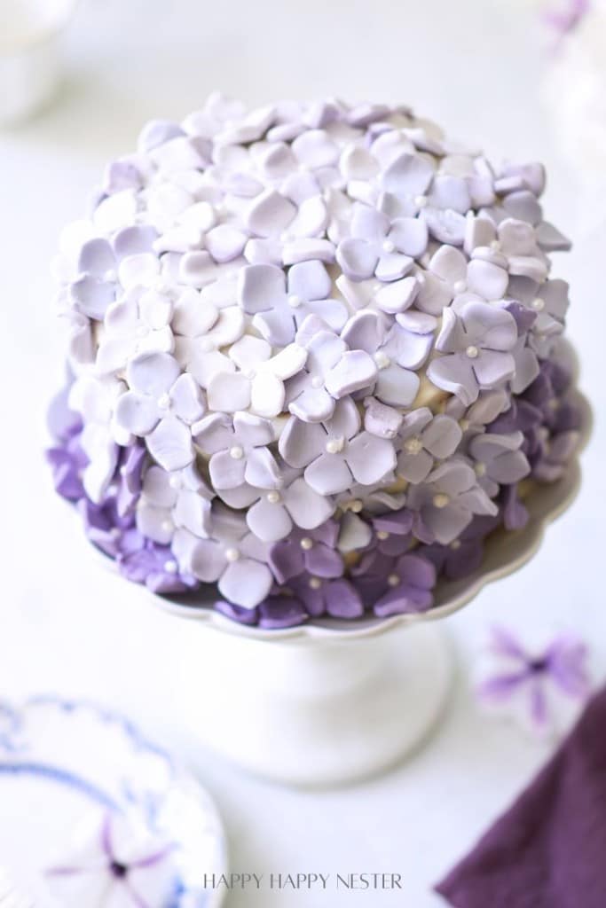 A beautifully decorated cake is covered in pastel purple and white flower-shaped icing. The cake is displayed on a white pedestal stand. The flowers create a gradient effect, starting with darker purple at the bottom and fading to lighter shades at the top.