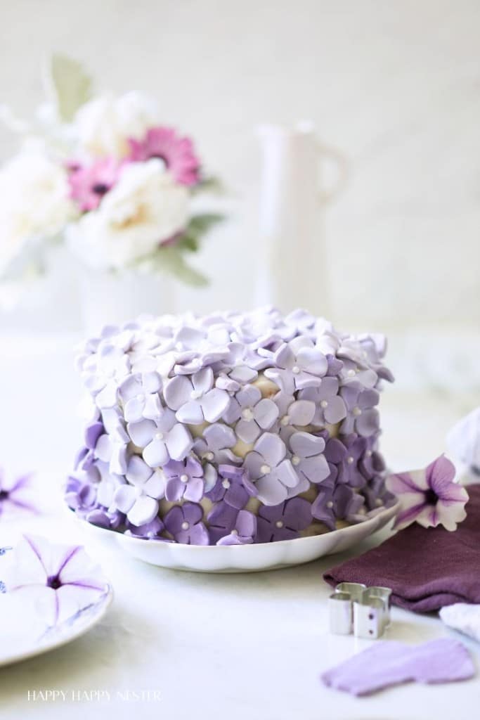 A beautifully decorated cake with purple and lavender flower-shaped icing sits on a white plate. In the background, a blurred vase with pink and white flowers and a white pitcher are visible. The cake is the focal point, conveying a delicate and elegant presentation.