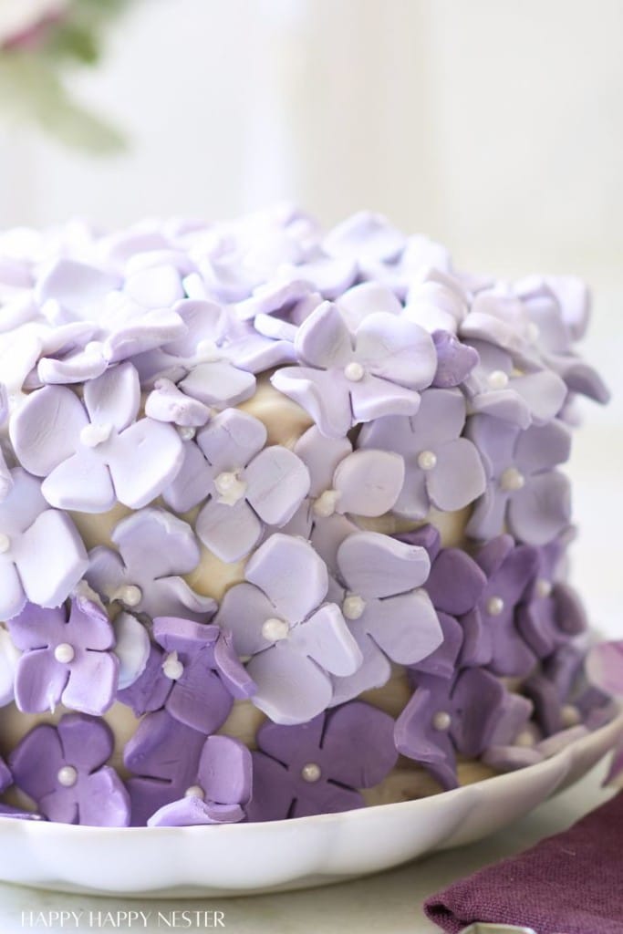 A beautifully decorated cake covered in pastel purple and white sugar flowers. The flowers are intricately arranged, creating a textured and elegant appearance. The cake sits on a white plate, and a folded purple napkin is partially visible in the foreground.