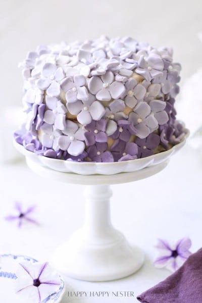 A beautifully decorated cake on a white cake stand. The cake is adorned with purple and lavender fondant flowers, creating an elegant and intricate floral design. The background is light and blurred, making the cake the focal point of the image.