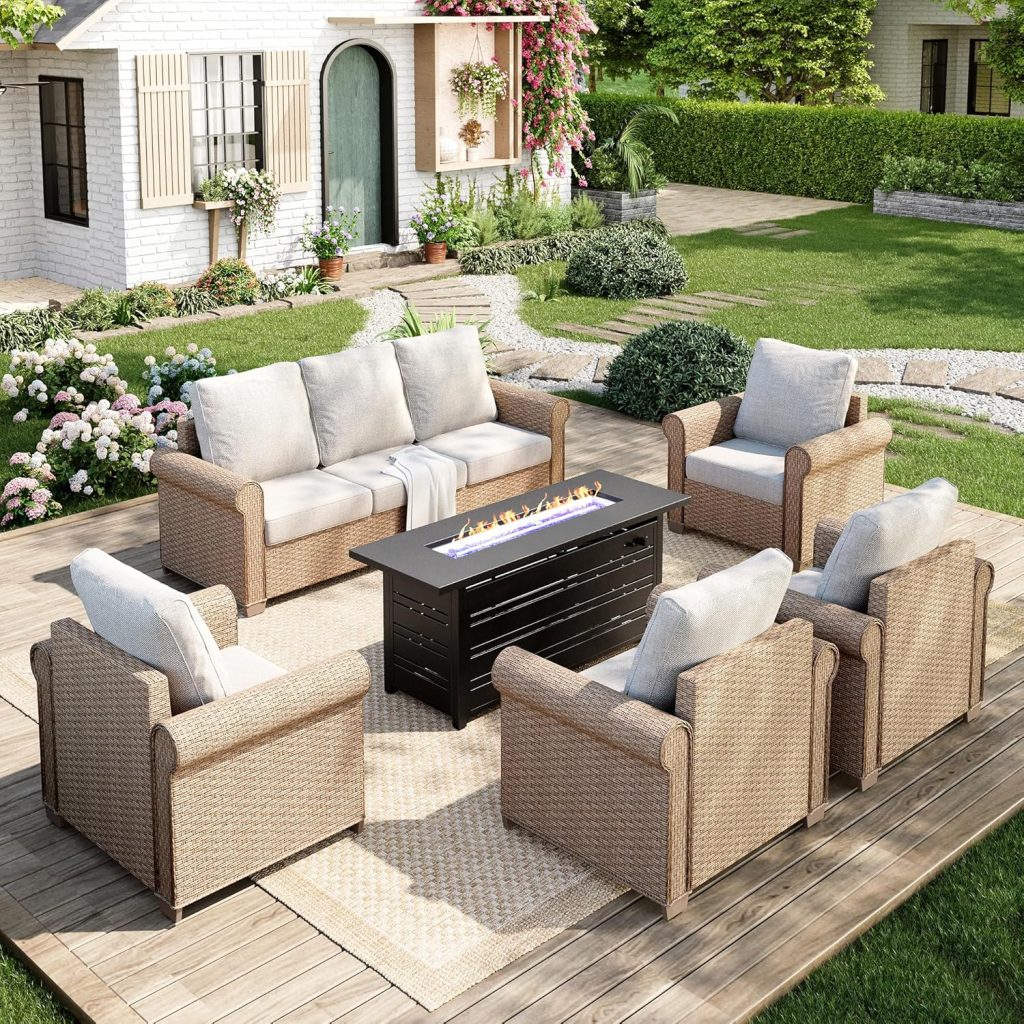 A cozy outdoor patio setup with wicker sofas and armchairs arranged around a rectangular fire pit table showcases inviting rattan decor ideas, placed on a wooden deck in a landscaped garden near a white house.